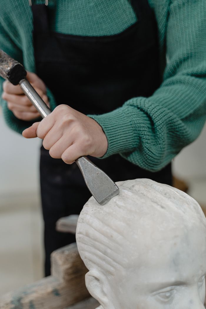 about-img A sculptor skillfully uses a chisel to shape a marble statue indoors, showcasing artistry and craftsmanship.